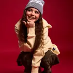 ZAH1DE mit langen braunen Haaren und einem Lächeln, trägt eine beige Sweatshirt, braune Hose und eine graue Mütze mit weißen Buchstaben, sitzt in der Hocke auf rotem Hintergrund.