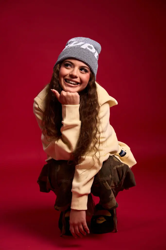 ZAH1DE mit langen braunen Haaren und einem Lächeln, trägt eine beige Sweatshirt, braune Hose und eine graue Mütze mit weißen Buchstaben, sitzt in der Hocke auf rotem Hintergrund.