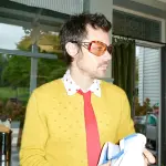 Harry Styles mit gelbem Pullover und roter Krawatte hält ein blaues und ein weiße Tuch in der Hand - Foto Credits: Johnny Dufort