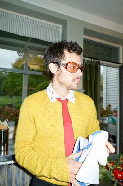 Harry Styles mit gelbem Pullover und roter Krawatte hält ein blaues und ein weiße Tuch in der Hand - Foto Credits: Johnny Dufort